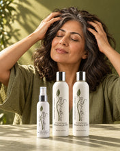 Load image into Gallery viewer, Woman with grey-streaked wavy hair massaging her scalp behind the three HSS Complete System bottles

