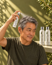 Load image into Gallery viewer, Man with short grey hair spraying the Bio Follicle Normal spray at his crown, shampoo and conditioner behind

